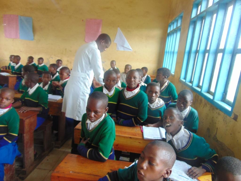 A health worker in a white coat administering deworming medicine to children in green school uniforms seated at their classroom desks in Njombe