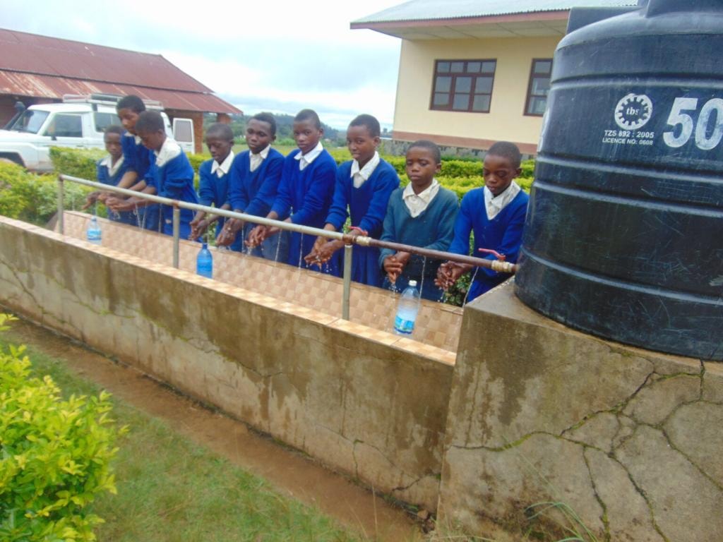 A row of older pupils in blue school uniforms washing their hands at a tiled outdoor basin fed by a 500-litre water tank at their school in Njombe
