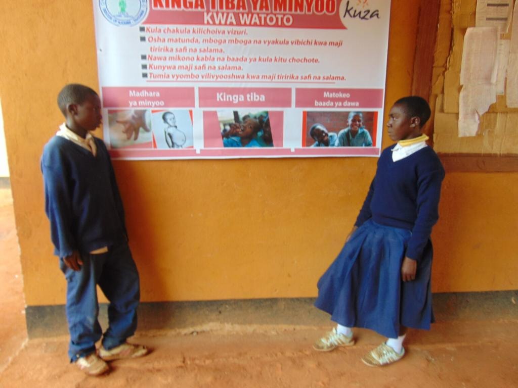 Two school pupils standing beside a Kuza deworming awareness poster reading 'Kinga Tiba ya Minyoo kwa Watoto' with the Kuza logo