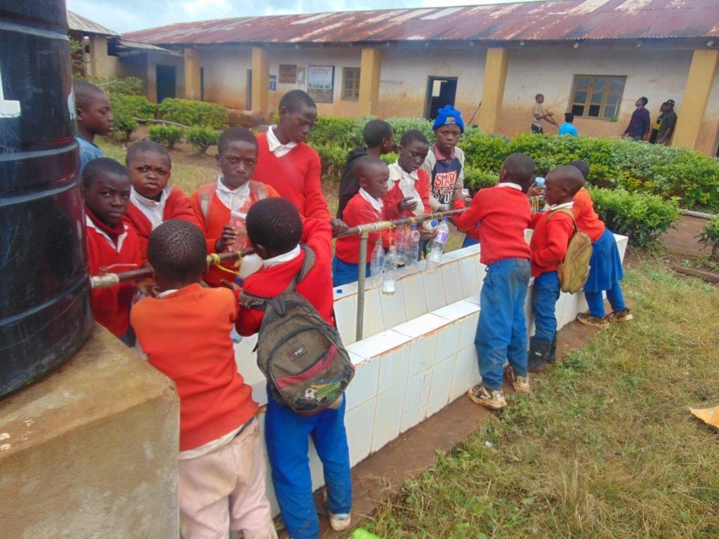 Children in red school uniforms collecting clean water from outdoor taps connected to a large black storage tank at their school in Njombe