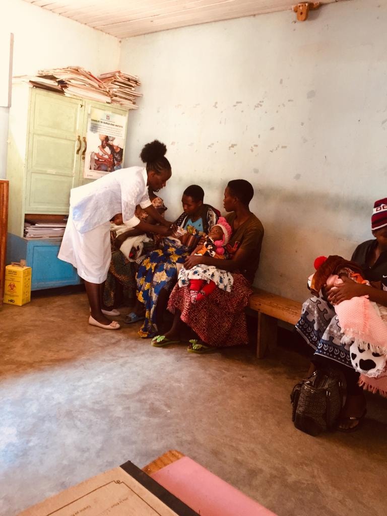 A nurse in white coat attending to a mother and baby while other mothers with infants wait at the RCH clinic in Njombe