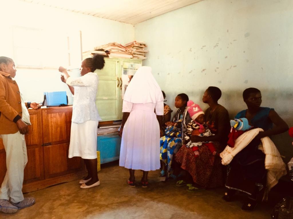 A Catholic sister, nurse, male visitor and mothers with children gathered in the RCH clinic room in Njombe — showing the Caritas Diocese partnership